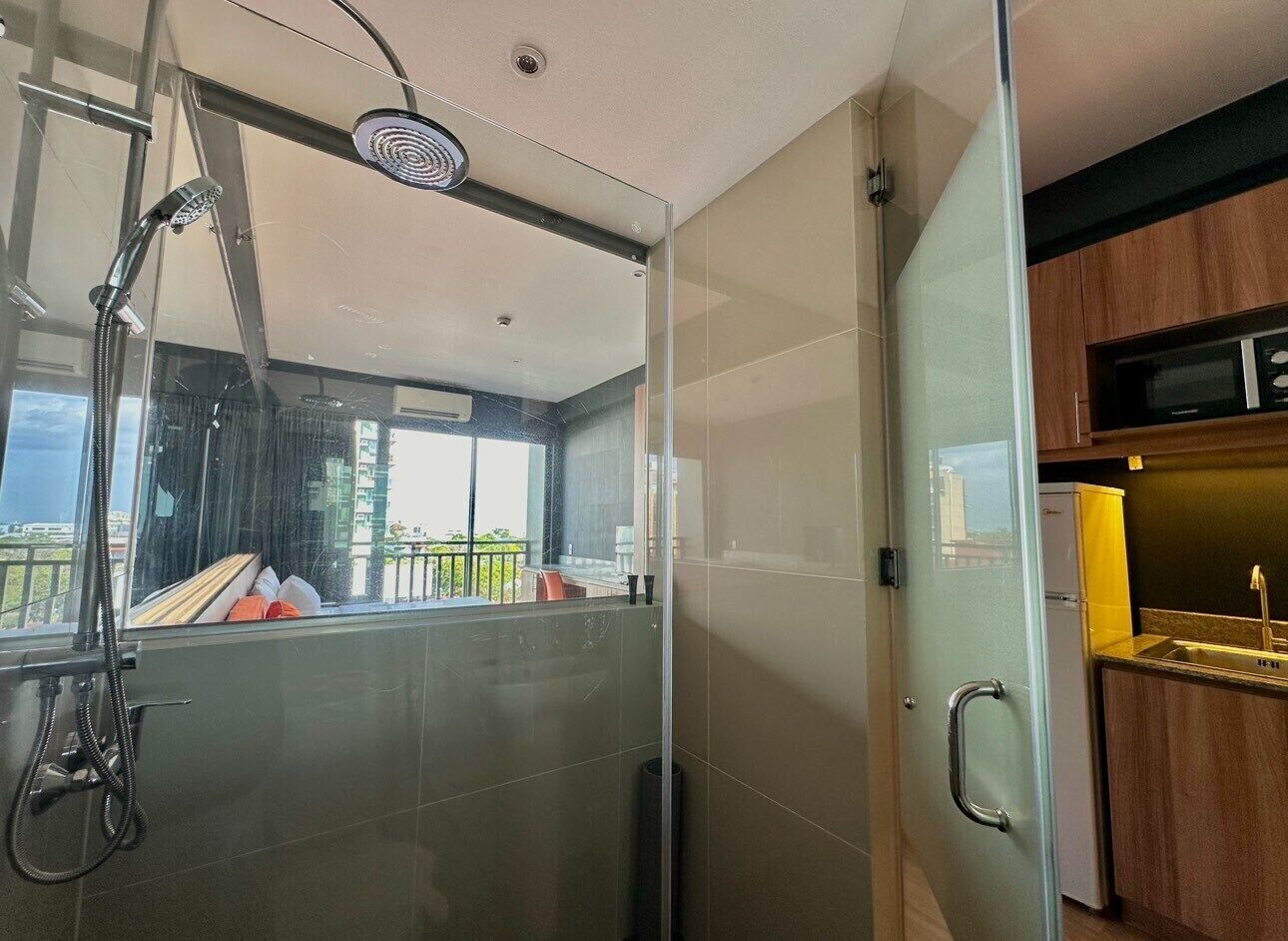 A modern bathroom with a glass shower enclosure, a visible balcony, and a kitchen area with wooden cabinets.