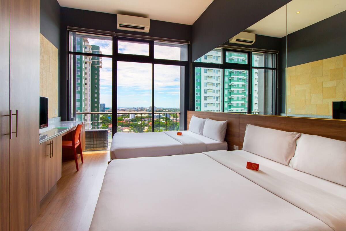 A hotel room with two beds, a desk, and a large window offering a city view.
