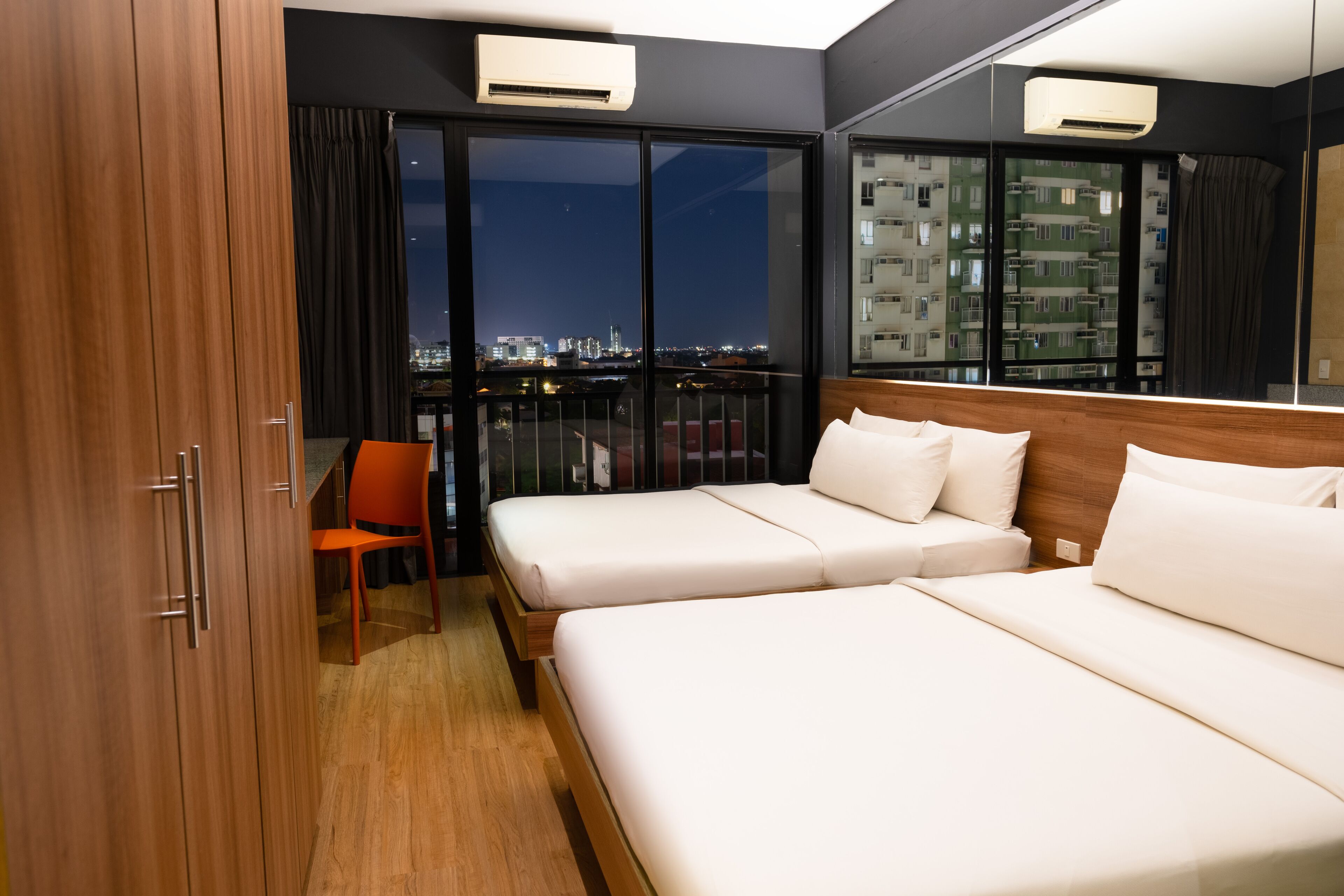 Habitación de hotel con dos camas, una silla y un amplio ventanal con vista a la ciudad.