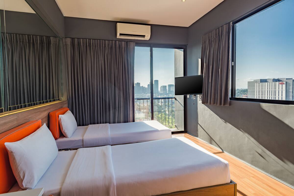 A modern hotel room with two beds, a large window with a city view, a flat-screen TV, and an air conditioning unit.