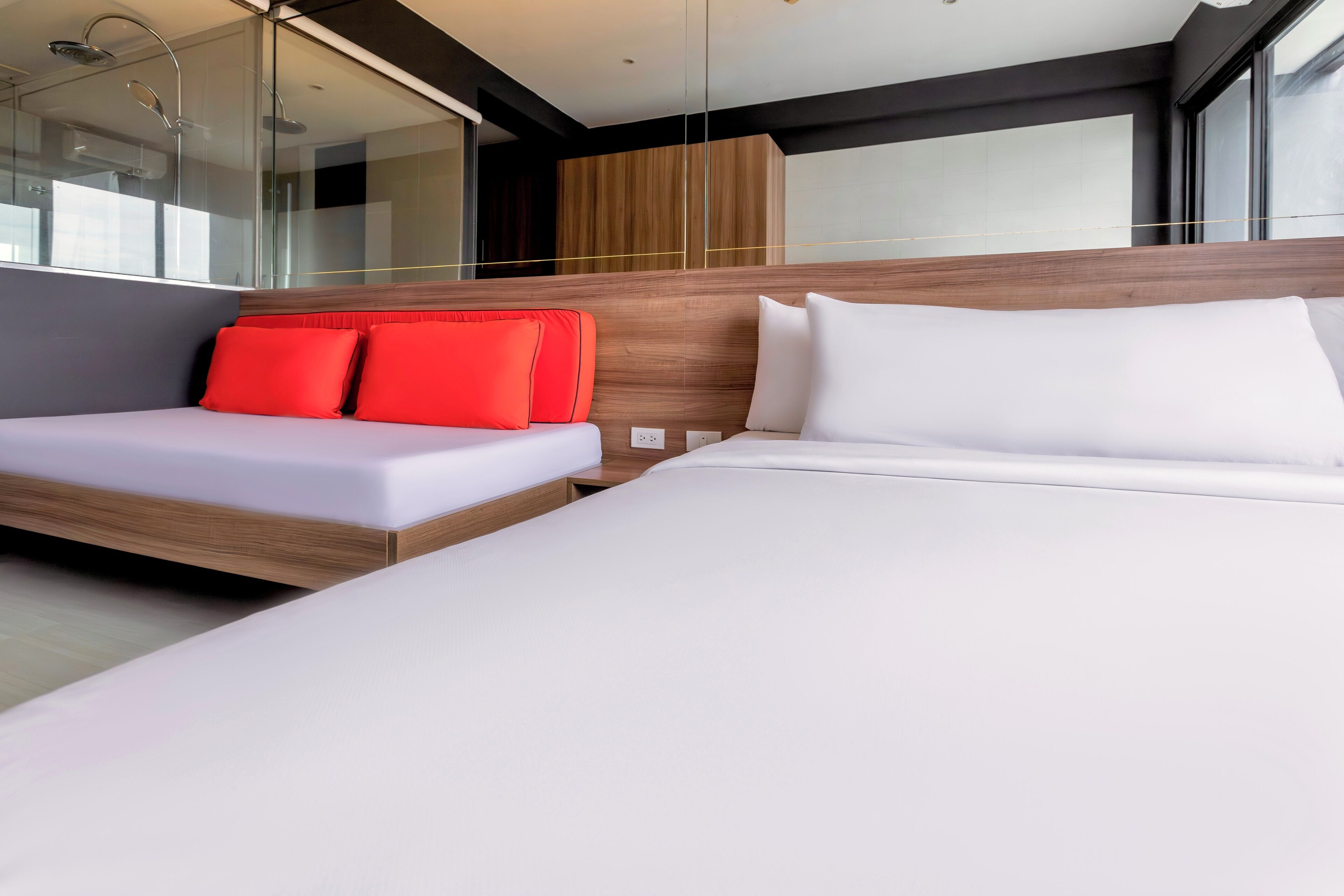 A modern hotel room with a large bed, a sofa with red cushions, and a glass shower enclosure.