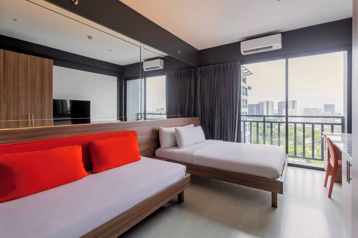 A modern hotel room with a large bed, a sofa with red cushions, a TV, and a balcony with a city view.