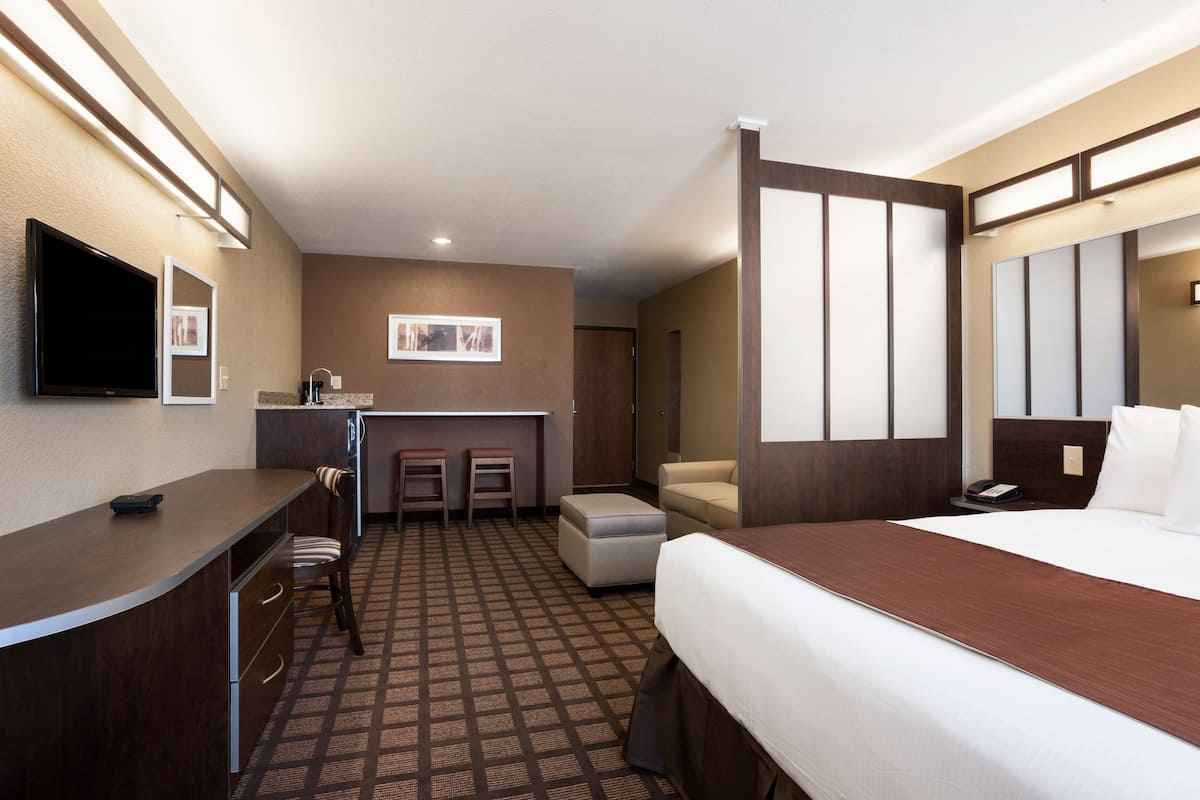 A hotel room with a bed, a desk, a chair, a small kitchenette, and a TV.