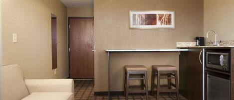 A compact hotel room with a kitchenette, a sofa, two bar stools, and a framed picture on the wall.