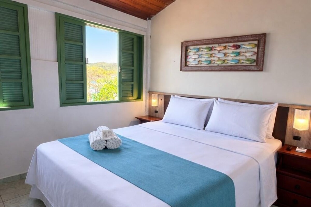 A neatly made bed with white and teal bedding, a wall-mounted fish painting, and a window with green shutters.