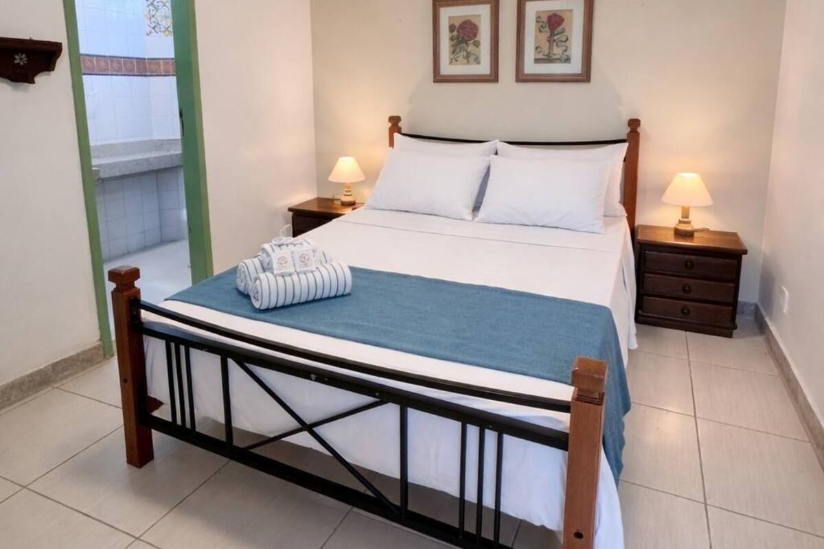 A bedroom with a bed, two bedside tables, a nightstand, a window with a green frame, and two wall-mounted pictures.