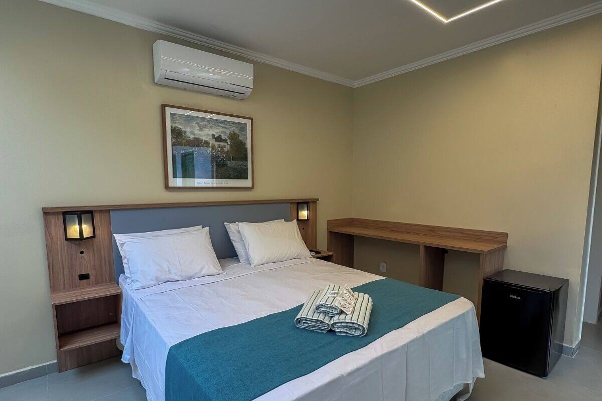A neatly made bed with a teal blanket, a framed picture above it, and a small bedside table with a lamp.