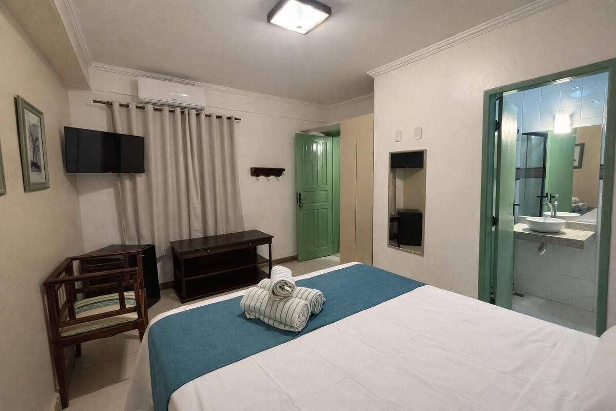 A hotel room with a bed, a television, a desk, a wardrobe, and a bathroom with a mirror and sink.