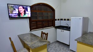 A small kitchen with a TV mounted on the wall, a window with wooden shutters, a sink, and a refrigerator. - Hotel da Canoa (Arraial do Cabo)