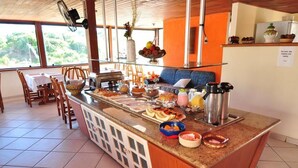 A breakfast bar with various food items, a seating area with chairs and a sofa, and a view of greenery through large windows. - Hotel da Canoa (Arraial do Cabo)