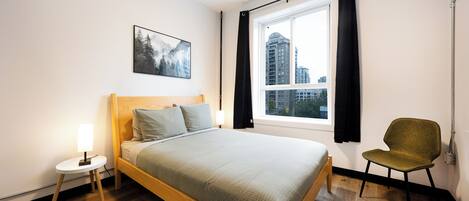 A modern bedroom with a wooden bed, a small bedside table, a chair, a window with curtains, and a cityscape view.