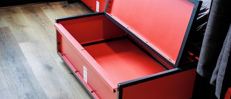 Een rode metalen opbergunit met een scharnierend deksel, staand op een houten vloer.