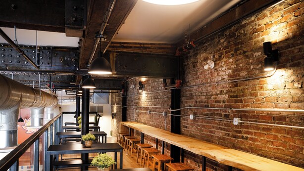 Un restaurant avec de longues tables et des bancs en bois, des murs en briques, des tuyaux apparents et un plafond haut avec des poutres métalliques.