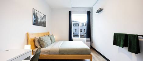 A modern bedroom with a wooden bed, a desk, and a view of buildings through a window.