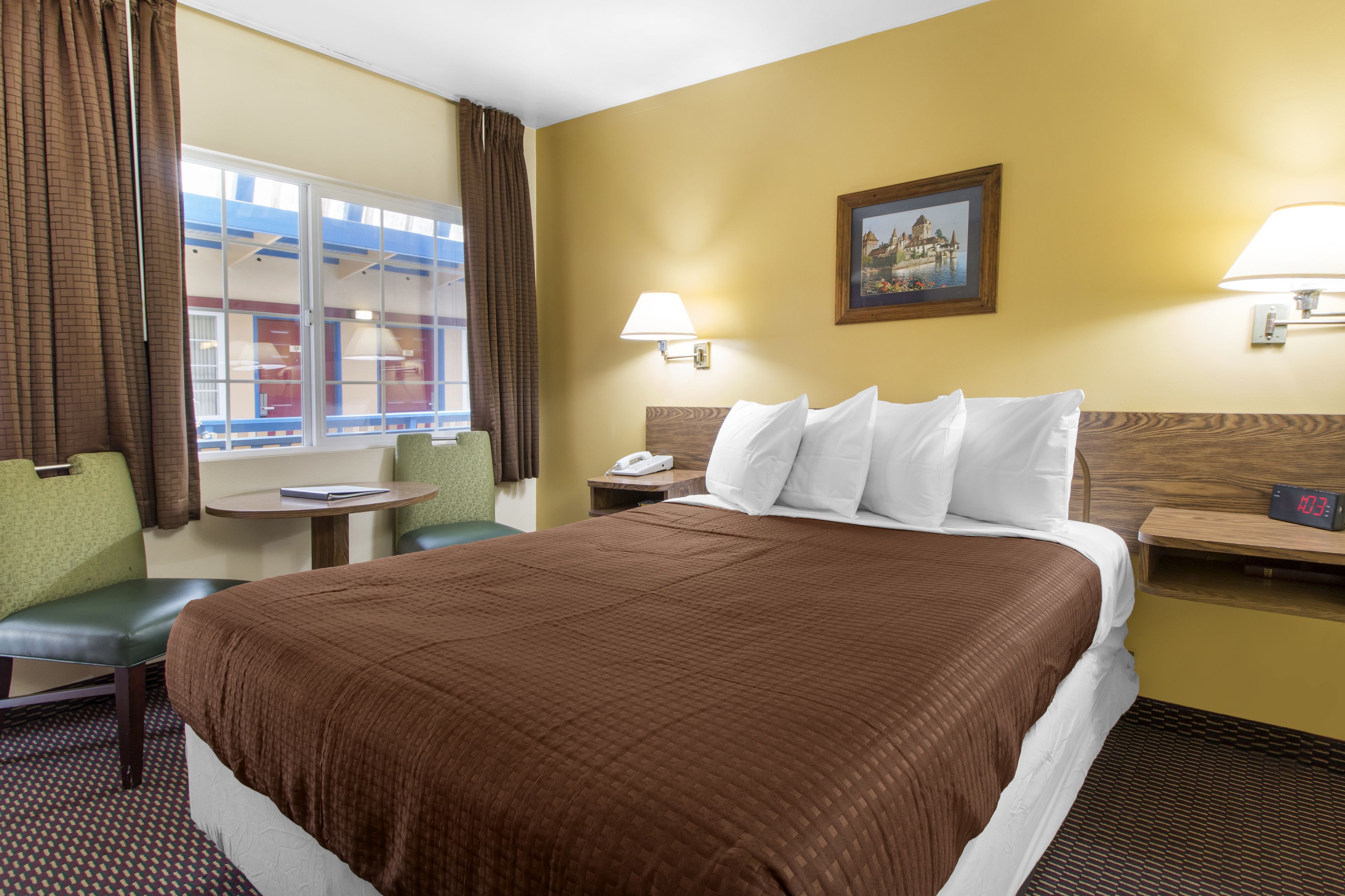 A hotel room with a large bed, a desk with a lamp, a chair, a window with curtains, and a painting on the wall.