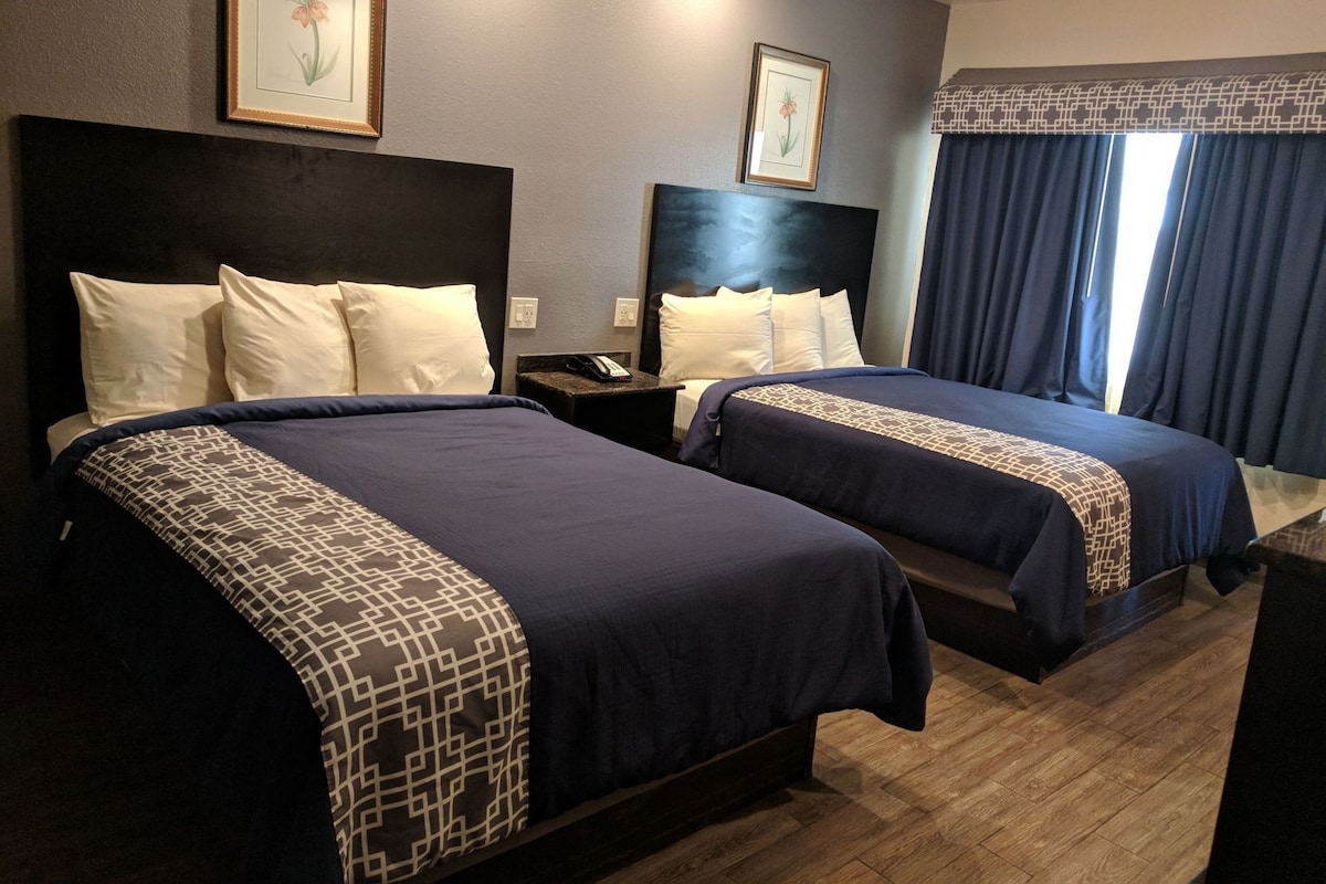 A hotel room with two beds, a dark headboard, a nightstand with a phone, and a window with curtains.