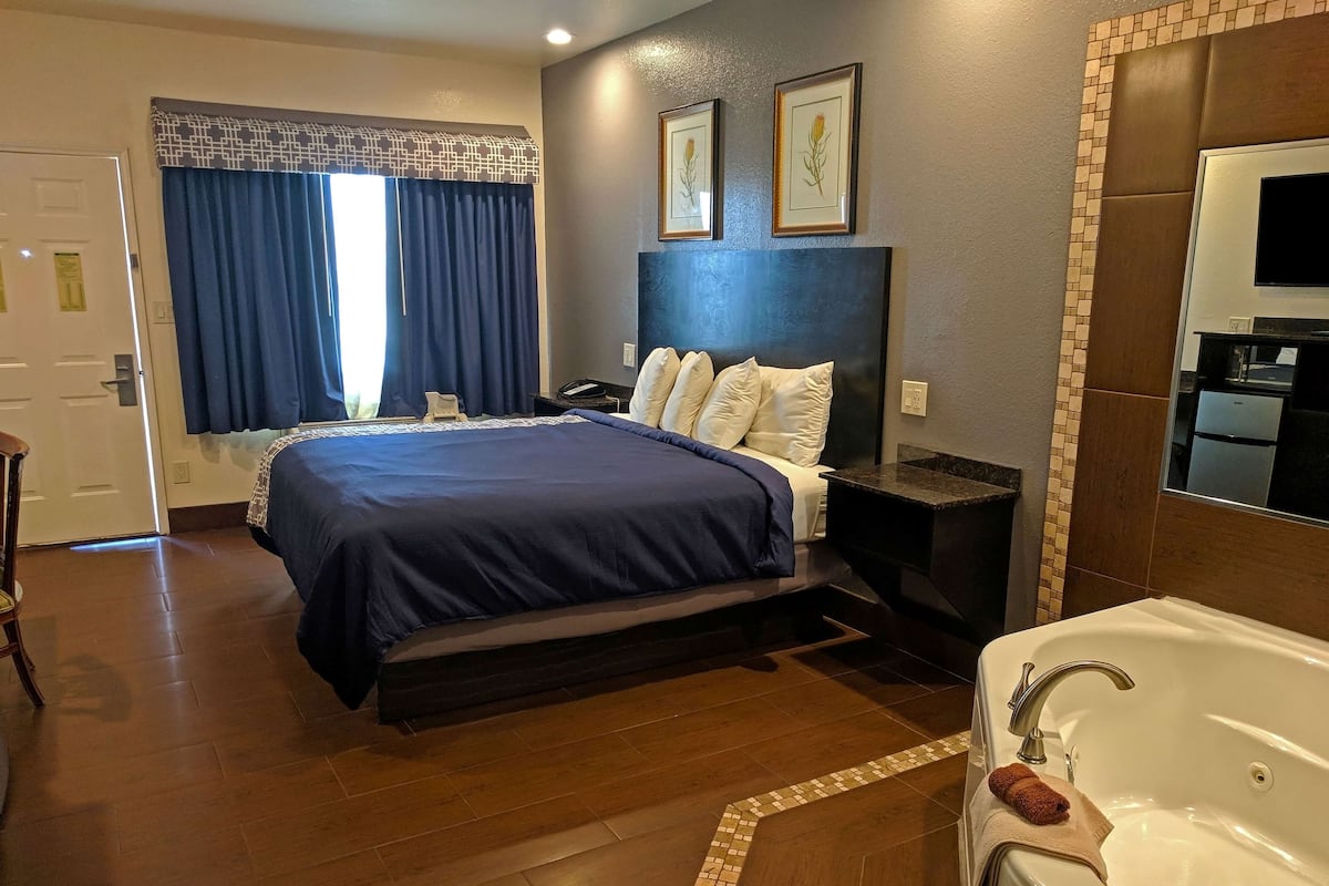 Une chambre d’hôtel avec un grand lit, une table de chevet, une commode, une salle de bain avec baignoire et une fenêtre avec des rideaux.