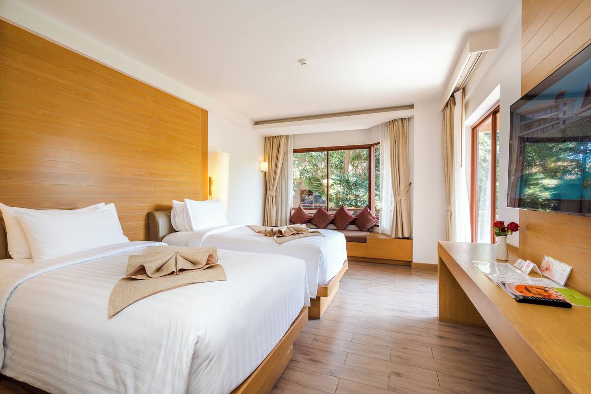 A hotel room with two beds, a wooden headboard, a large window with curtains, a wooden desk with a chair, and a television mounted on the wall.