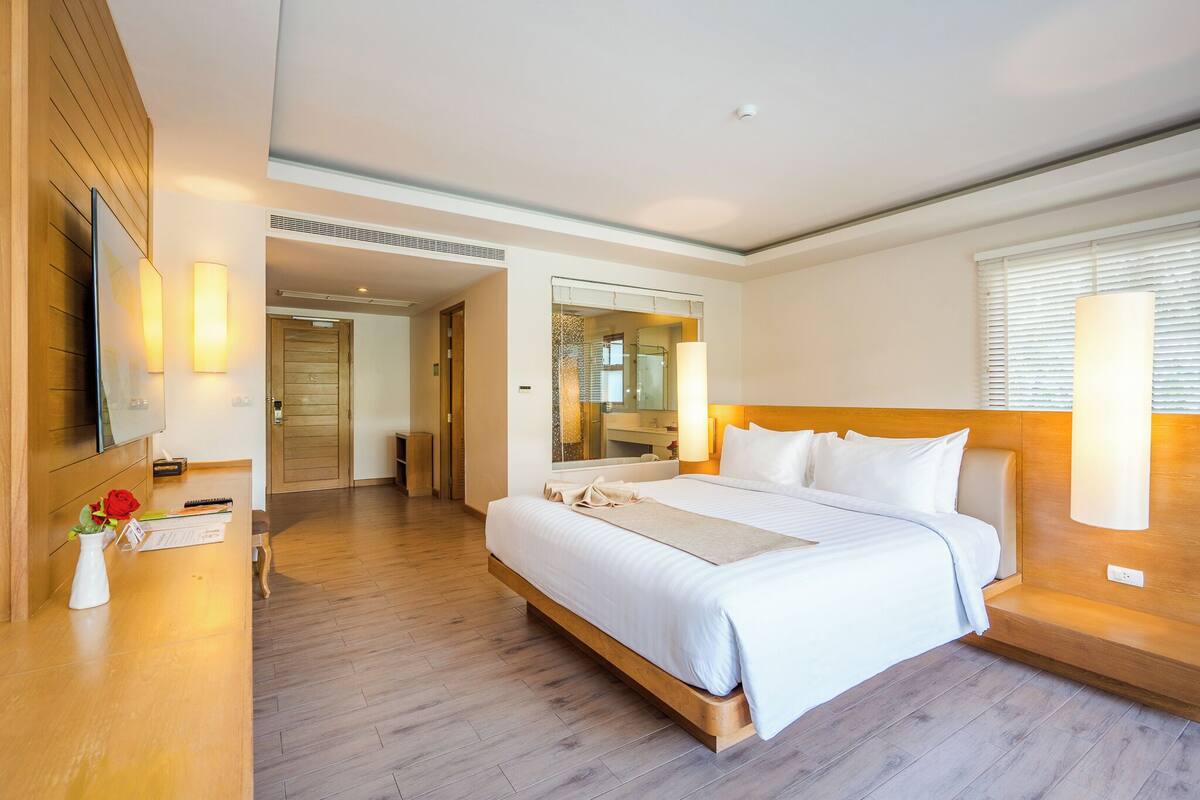 A hotel room with a large bed, wooden headboard, a flat-screen TV, a bedside table with a vase and a book, and a mirror above the bed.