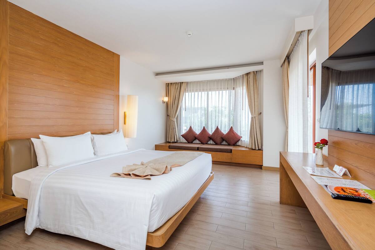A modern hotel room with a large bed, a wooden headboard, a seating area with cushions, a wooden desk, and a window with curtains.