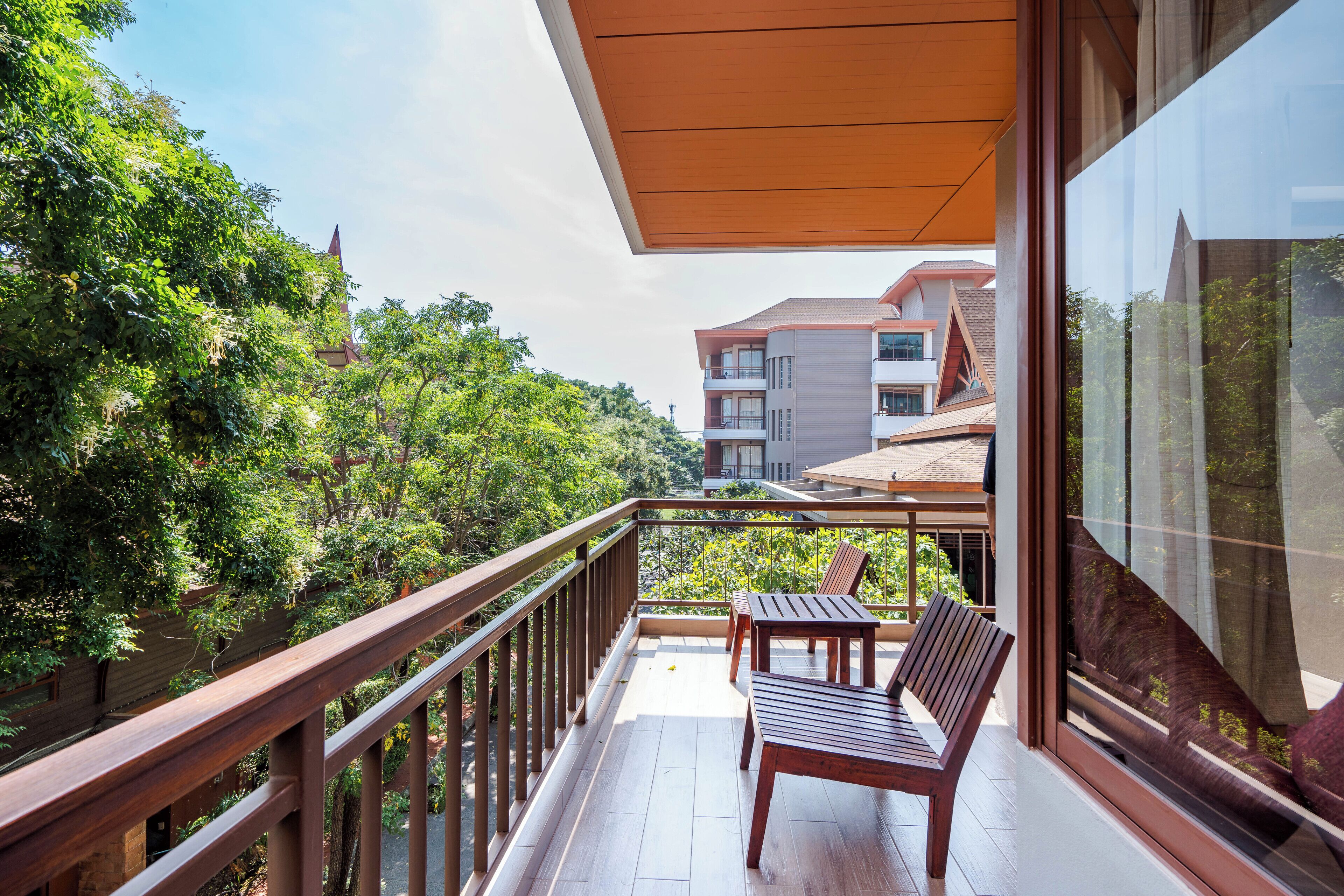 A balcony with wooden furniture, a glass railing, and a view of surrounding buildings and greenery.
