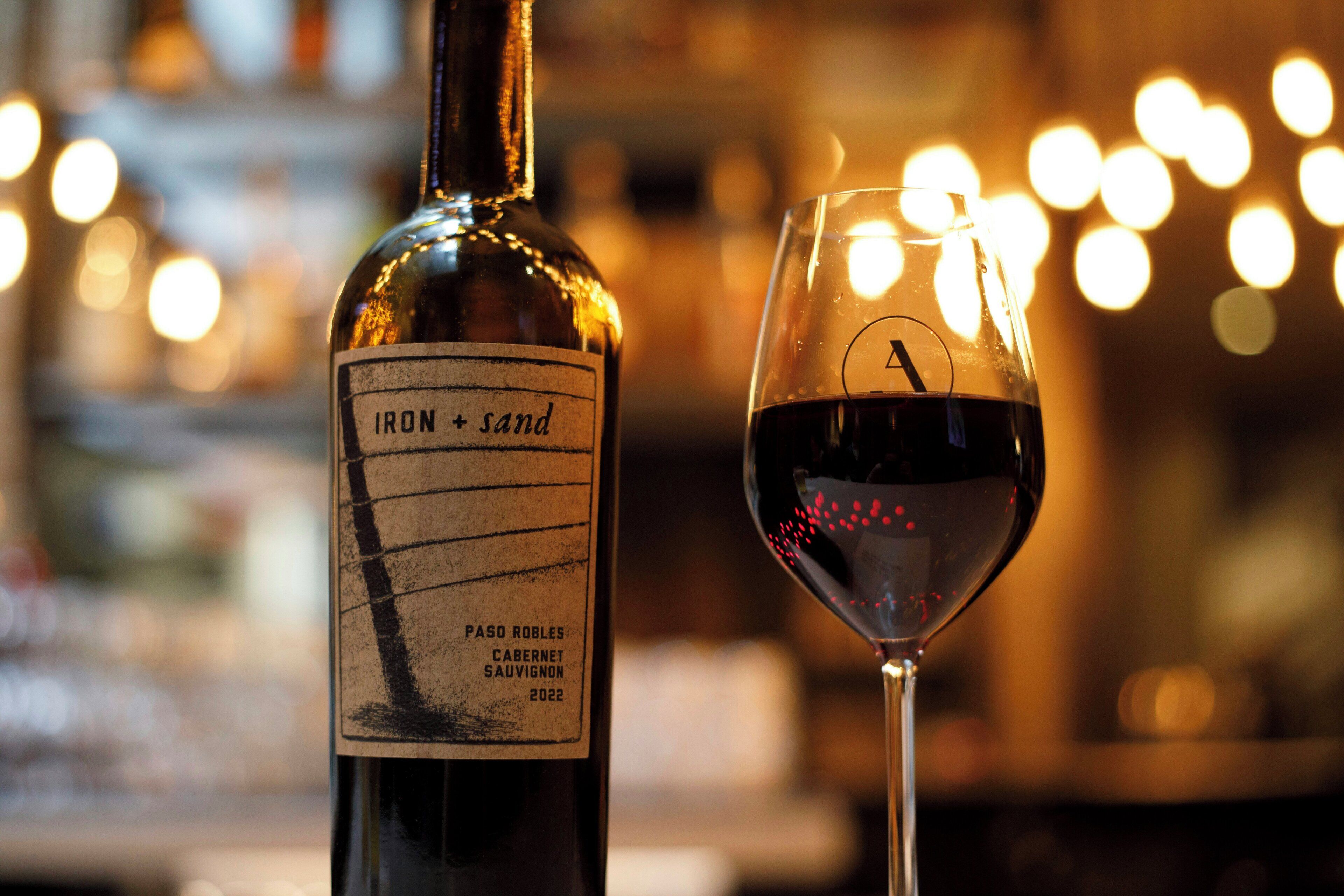 Eine Flasche Iron + Sand Paso Robles Cabernet Sauvignon 2022 und ein Weinglas mit Rotwein.
