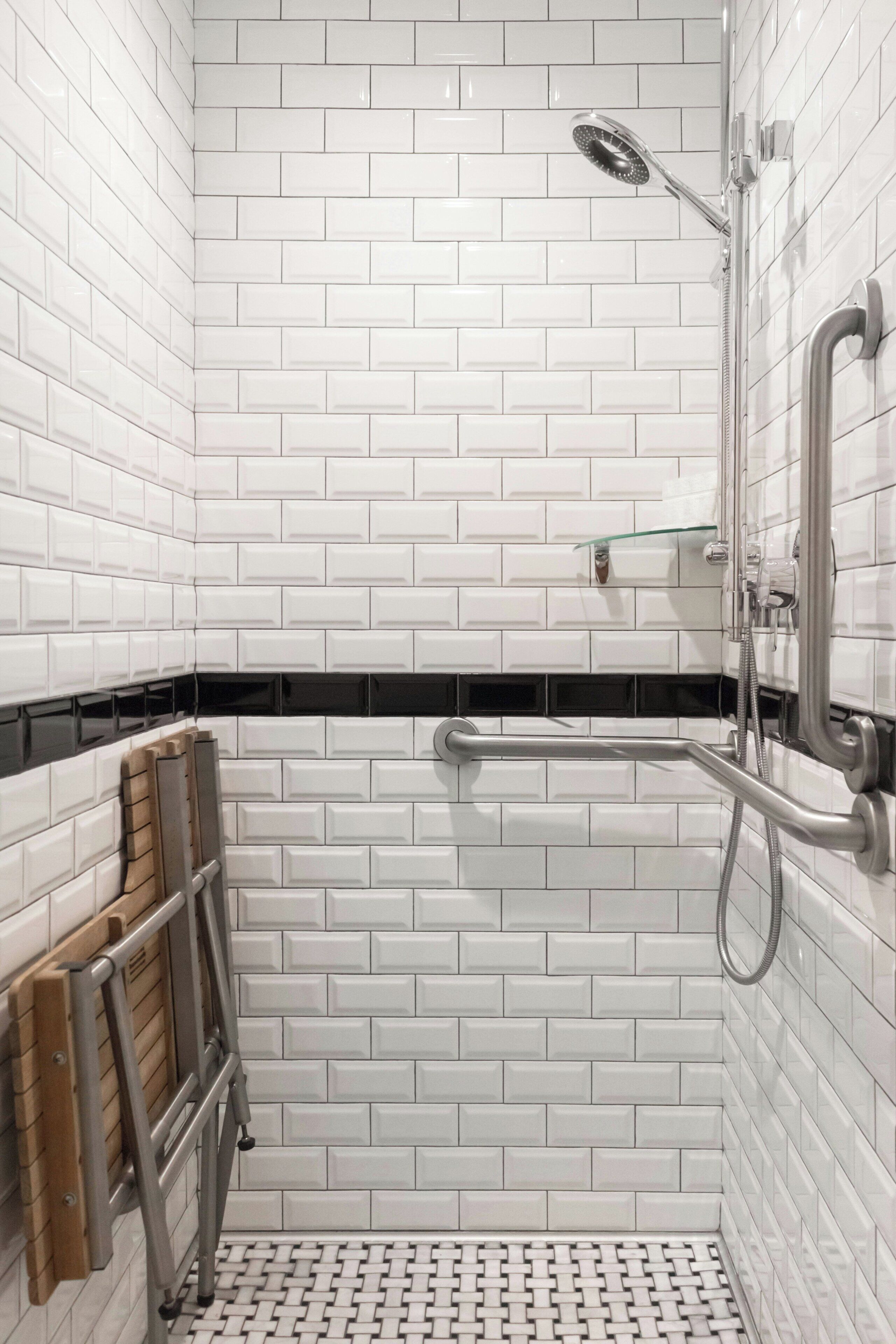 Zona de ducha con paredes de azulejos blancos, un estante de vidrio y una barra de apoyo de metal.