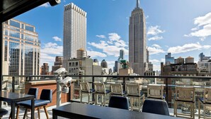 A rooftop terrace with a view of the Empire State Building, modern buildings, and a glass railing. - Archer Hotel New York (New York)