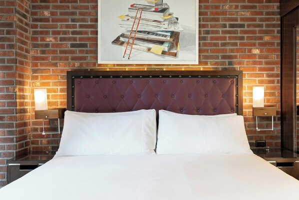 Habitación de hotel con pared de ladrillo, cabecero capitonado, cama con ropa blanca y dos lámparas de noche.