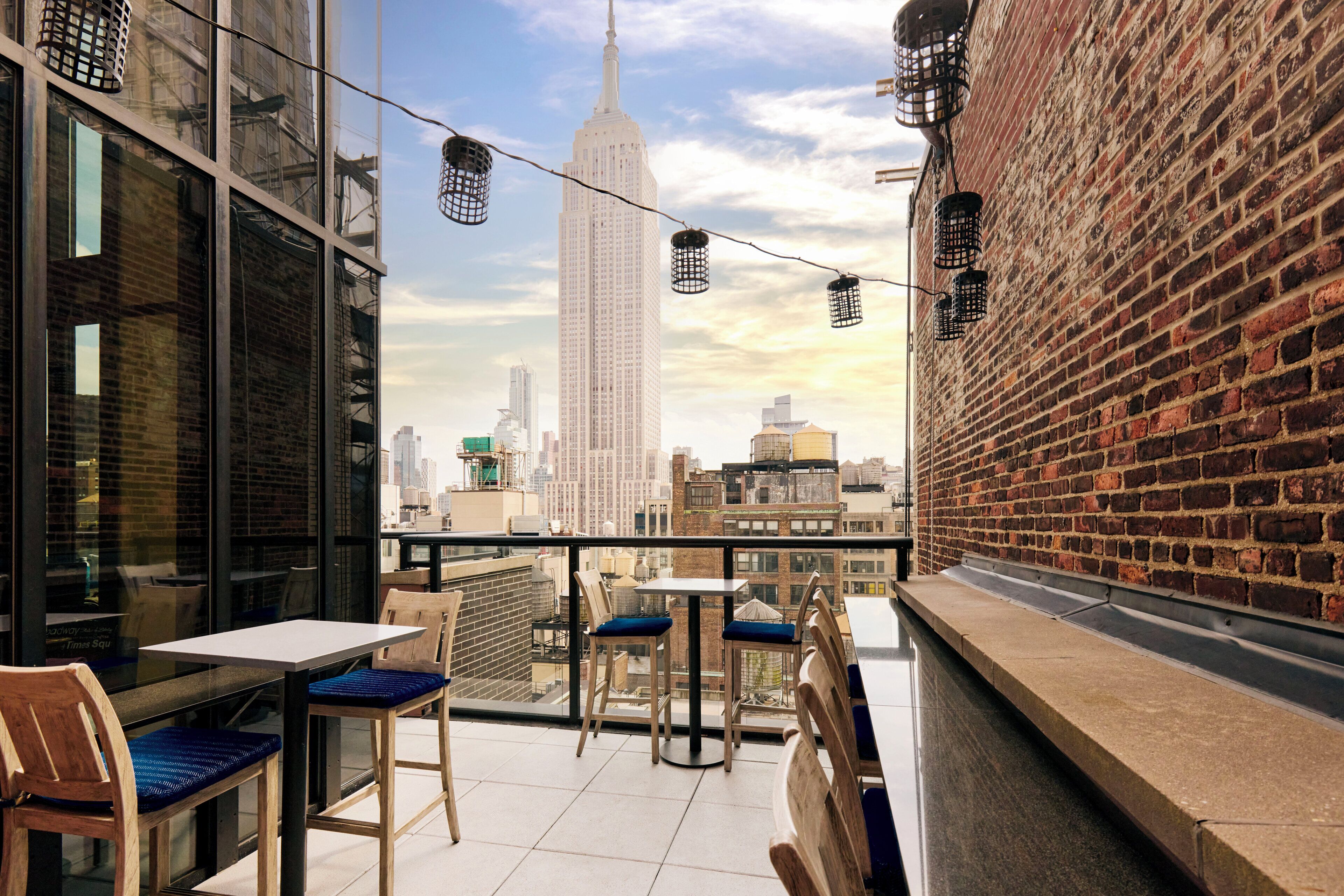 A rooftop terrace with tables and chairs, a brick wall, and a view of the Empire State Building.