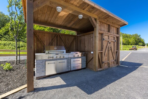 A covered outdoor kitchen with a stone countertop and stainless steel appliances, set in a wooden structure with a gravel floor.