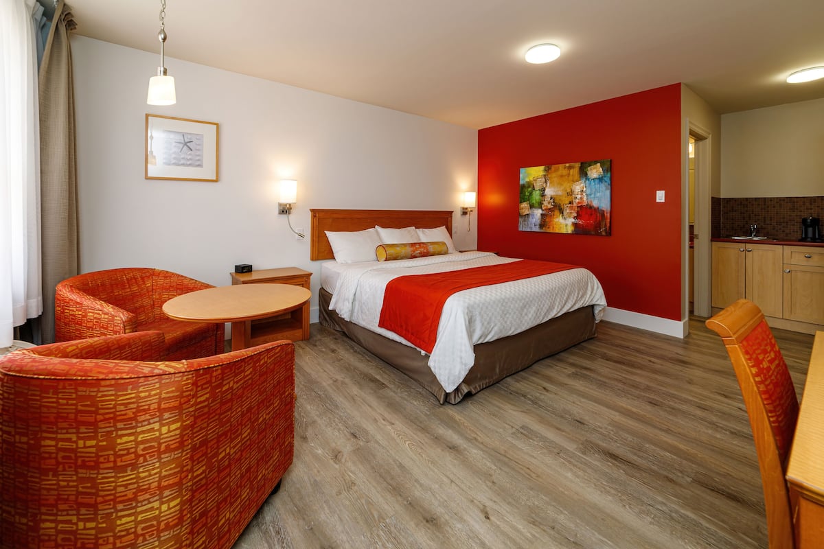 Ein Hotelzimmer mit einem Bett, zwei Stühlen, einem kleinen Tisch, einem Gemälde an der Wand und einer Küchenzeile.
