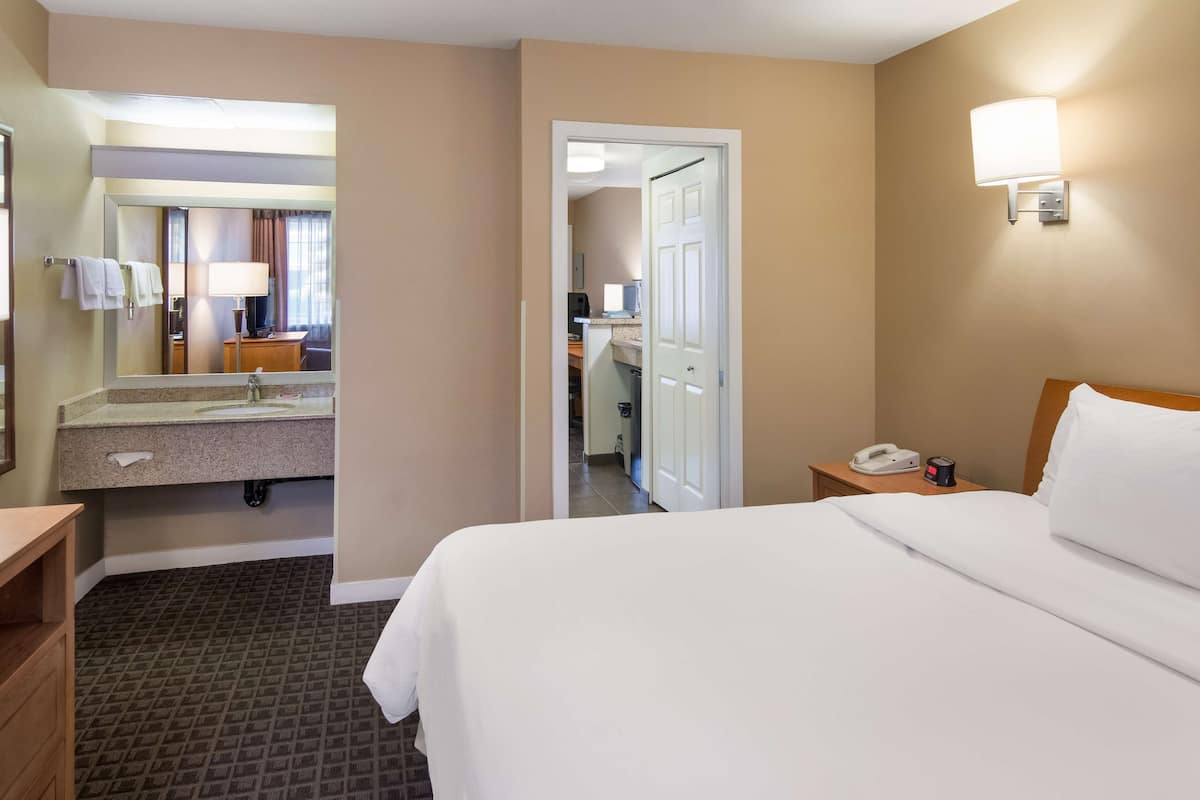 A hotel room with a bed, a bathroom with a sink and mirror, a bedside table with a phone, and a door leading to another room.