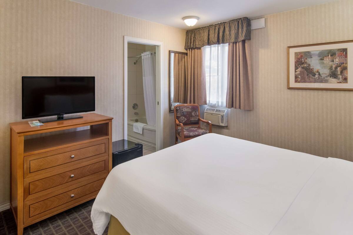 A hotel room with a bed, a TV on a wooden dresser, a chair, an air conditioning unit, and a painting on the wall.