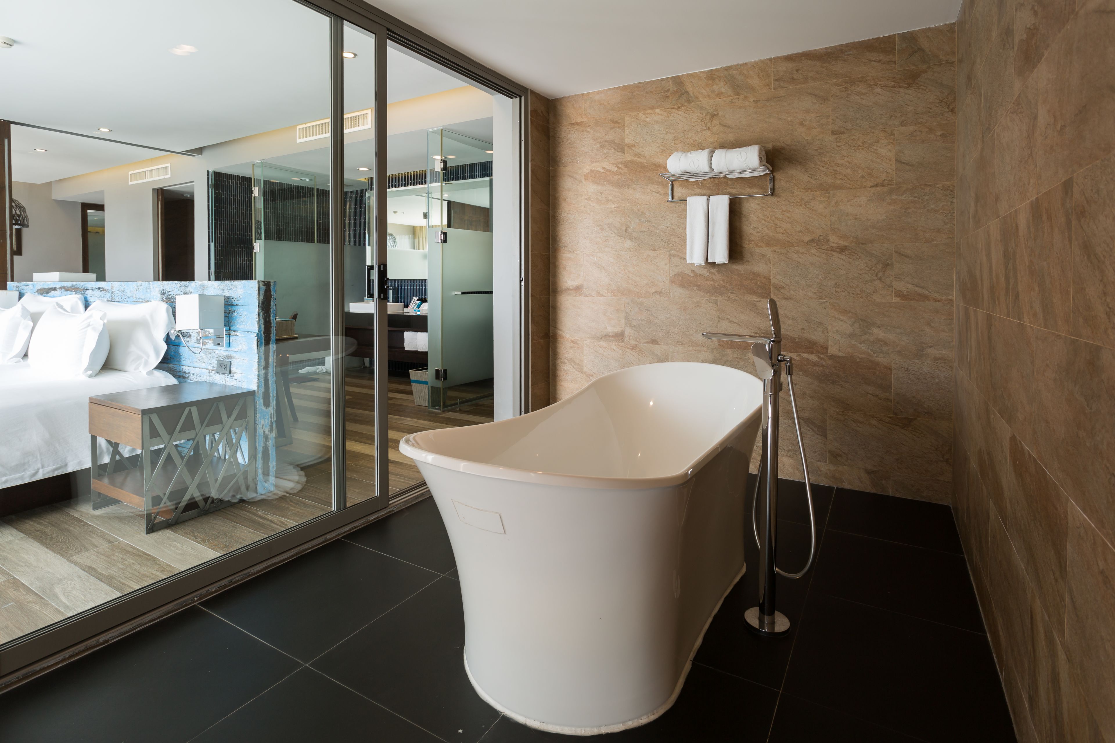 A modern bathroom with a freestanding bathtub, a glass shower enclosure, and a visible bedroom with a bed and a nightstand.