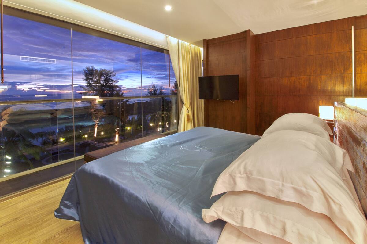 A modern hotel room with a large bed, a TV mounted on the wall, and a balcony with a view of the sunset.