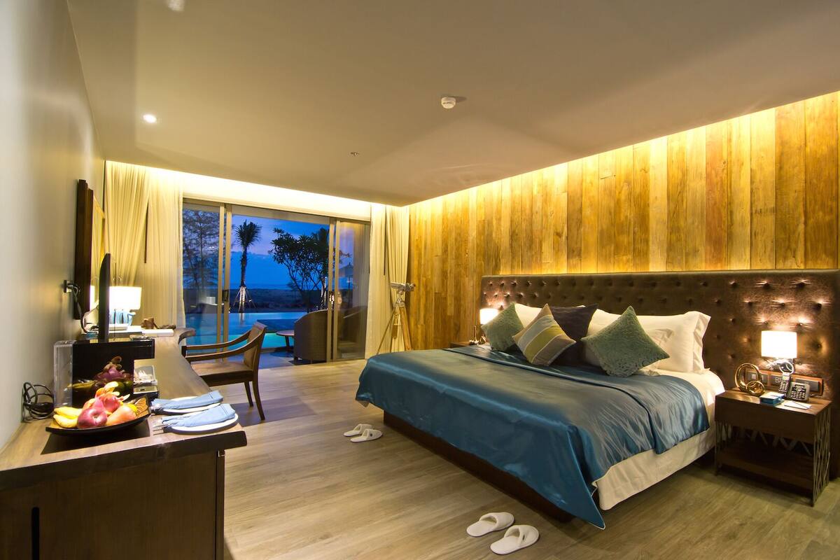 A hotel room with a bed, bedside table, and a desk with a computer. There is a view of the beach and palm trees outside the window.