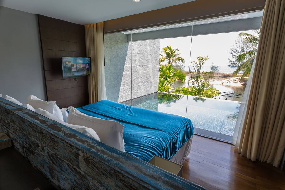 A modern hotel room with a large bed, a TV mounted on the wall, and a balcony with a view of a beach and palm trees.