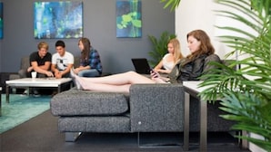 A modern lounge area with a grey sofa and armchairs, a coffee table, and a laptop on the sofa. There are abstract paintings on the wall and a plant in the foreground. - Griffith University Village (Southport)