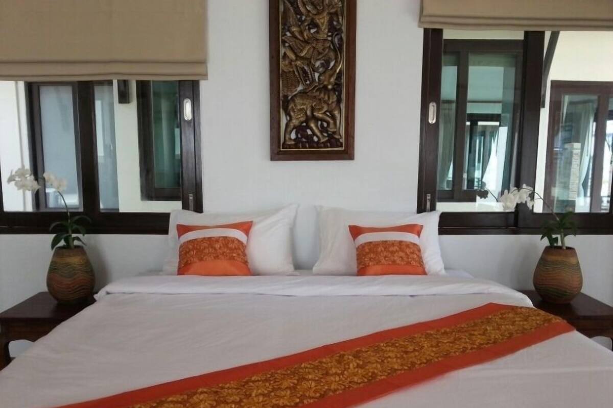 Una cama bien hecha con sábanas blancas y dos almohadas naranjas, flanqueada por dos mesitas de noche con jarrones decorativos.