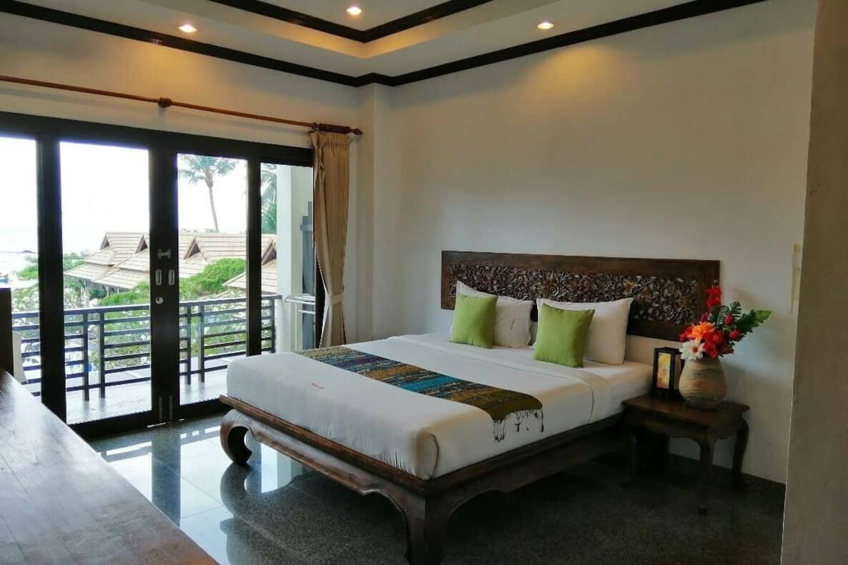 Un dormitorio con una cama grande, un cabecero de madera, una mesita de noche con un jarrón de flores y un balcón con vistas a edificios y zonas verdes.