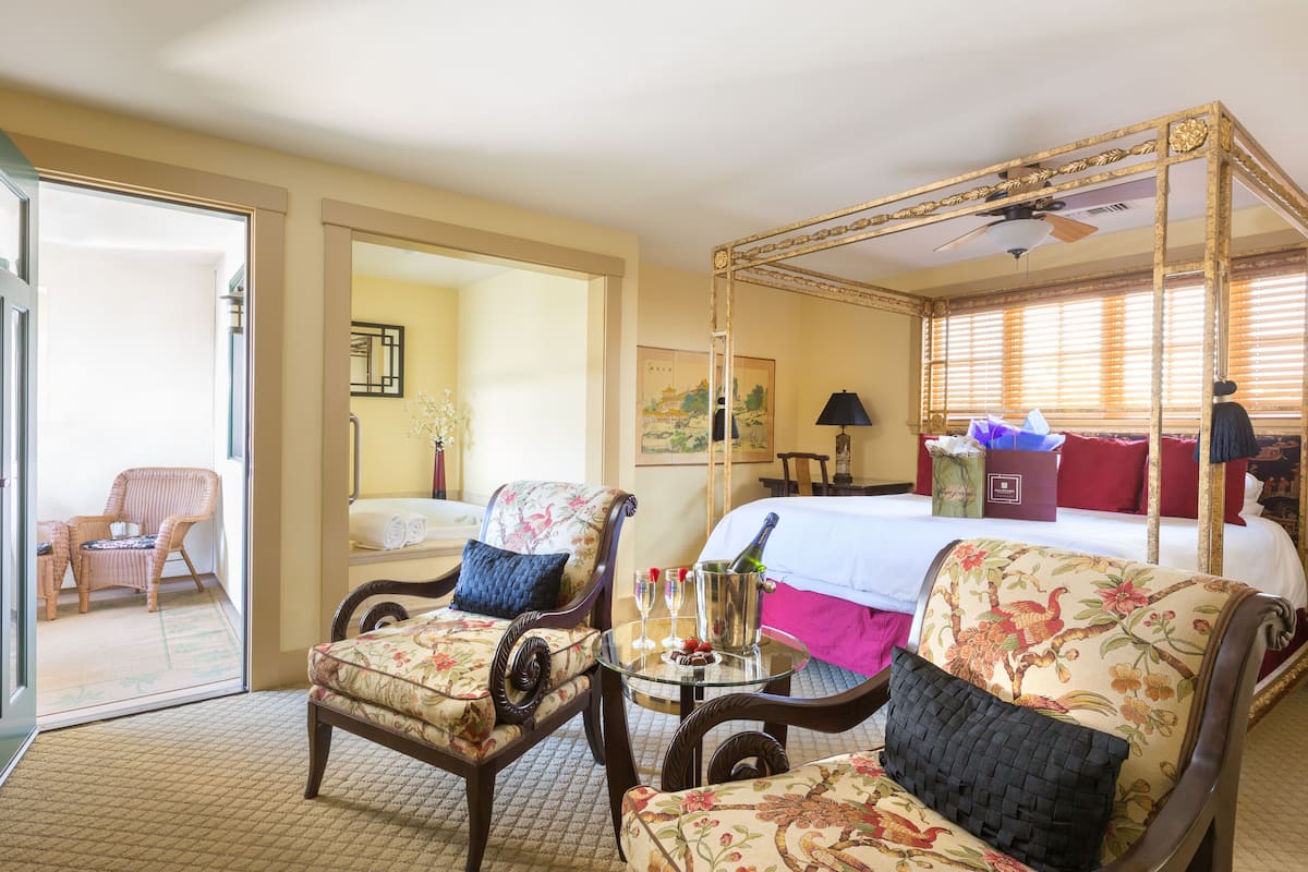 A hotel room with a four-poster bed, a sitting area with two chairs, a small table with a bottle of champagne, and a view of the outside through a door.