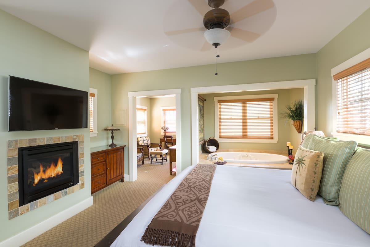 A bedroom with a fireplace, a large bed, a ceiling fan, and a view of a bathroom.