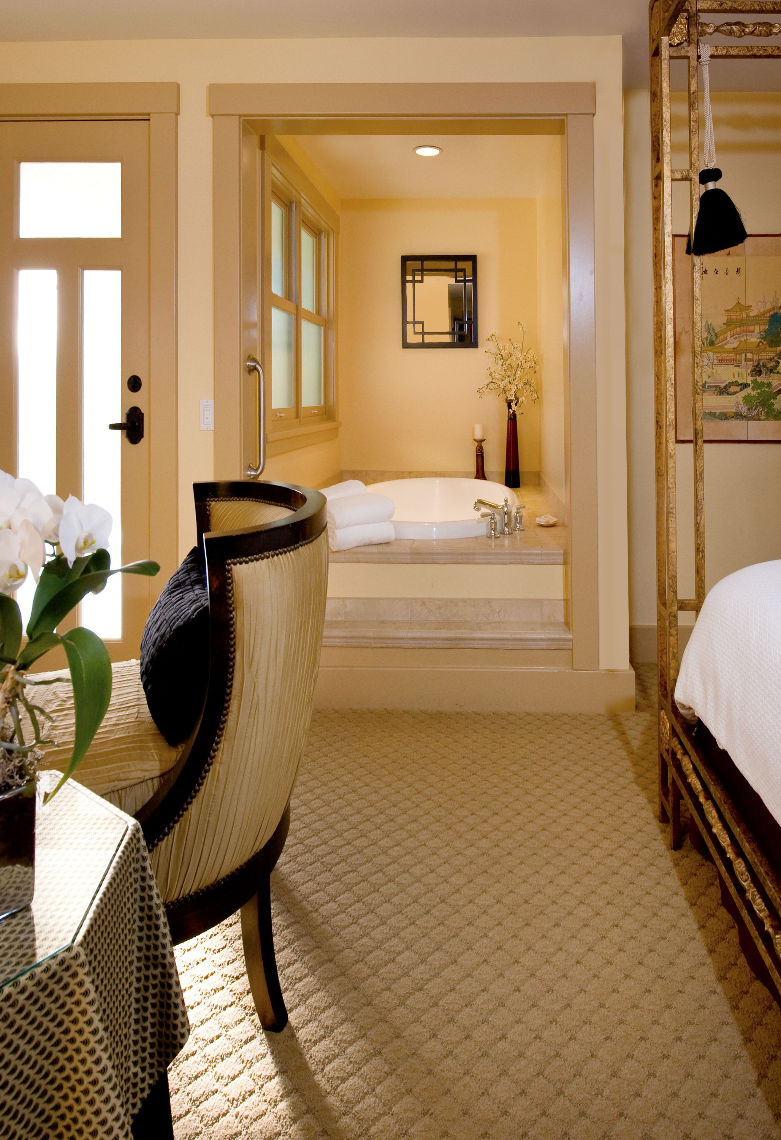 Ein Hotelzimmer mit einem großen Bett, einer Sitzecke inklusive Sessel, einem kleinen Tisch, einem Badezimmer mit Badewanne und einem Spiegel an der Wand.