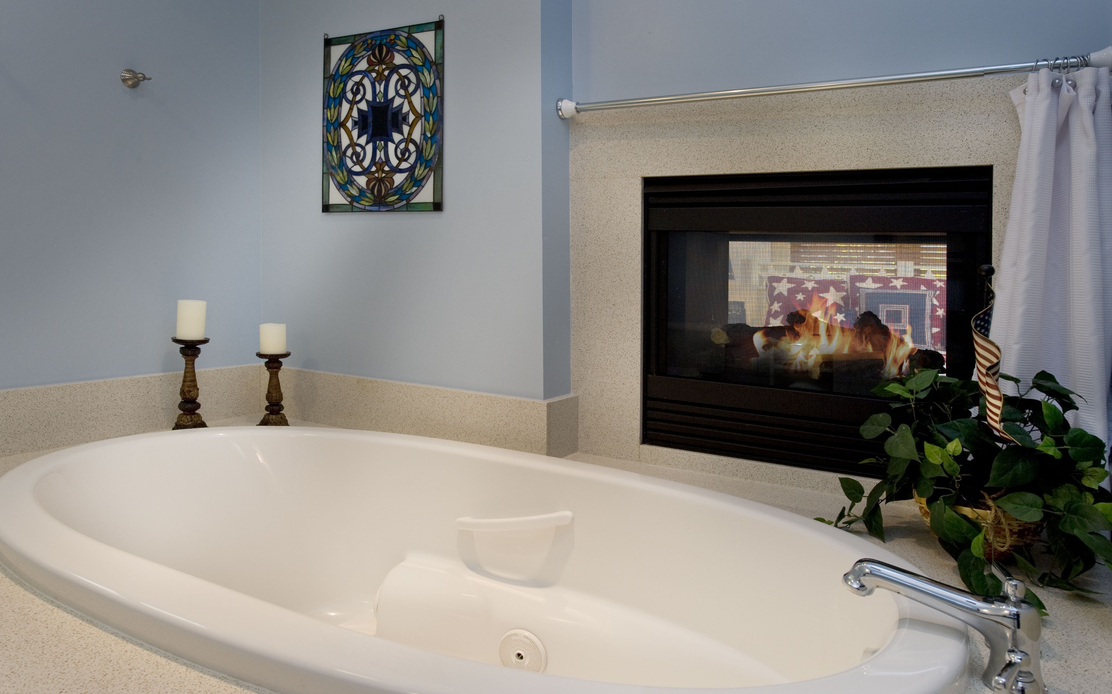 A bathroom with a bathtub, two candles on a stand, a decorative wall piece, and a fireplace with a cityscape view.