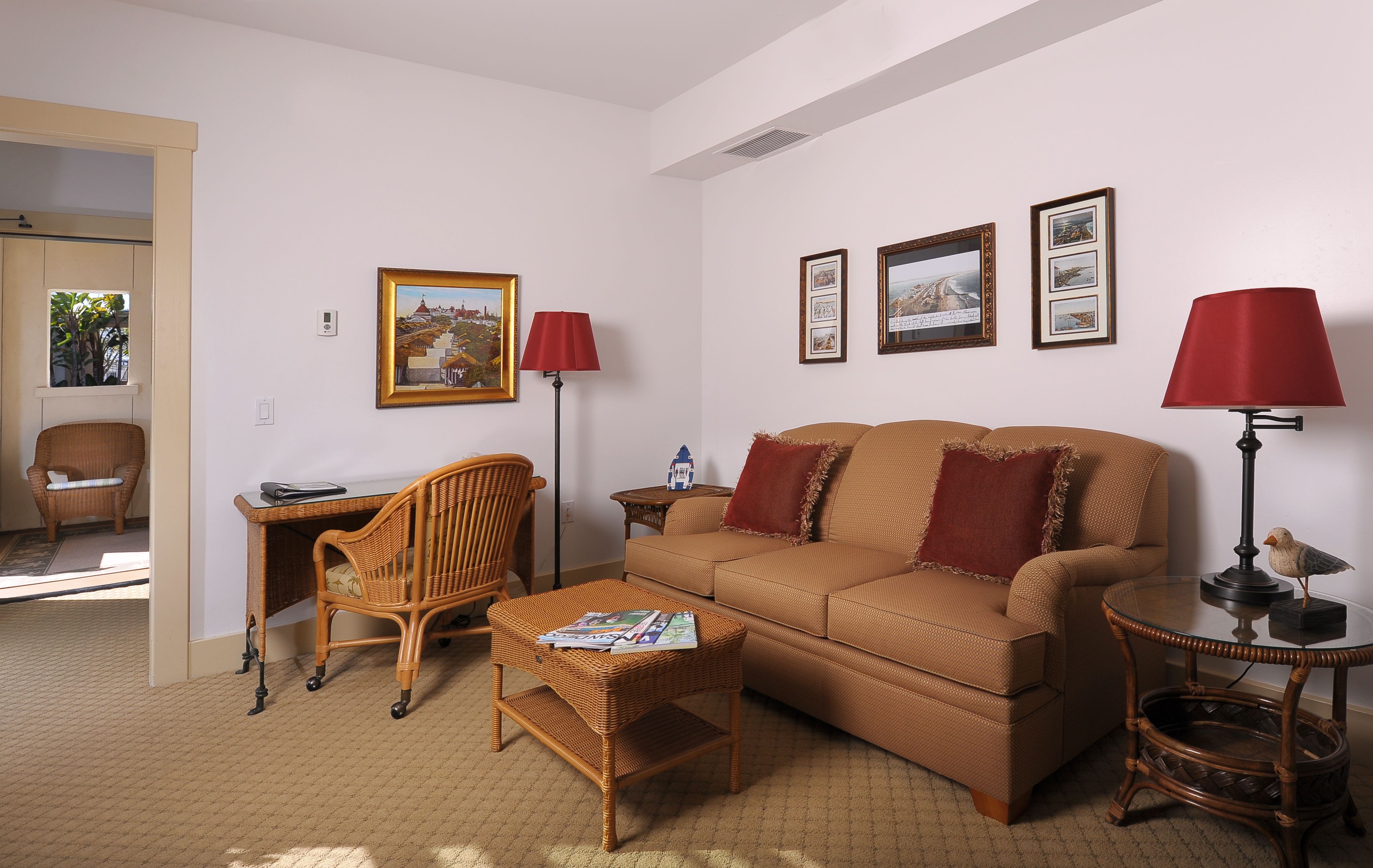 A living room with a brown sofa, a wicker coffee table, a wicker chair, a small side table, and a lamp. There are framed pictures on the wall.