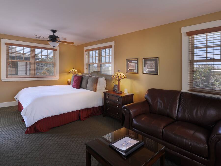 A hotel room with a bed, a leather sofa, a nightstand with lamps, and two windows with blinds.