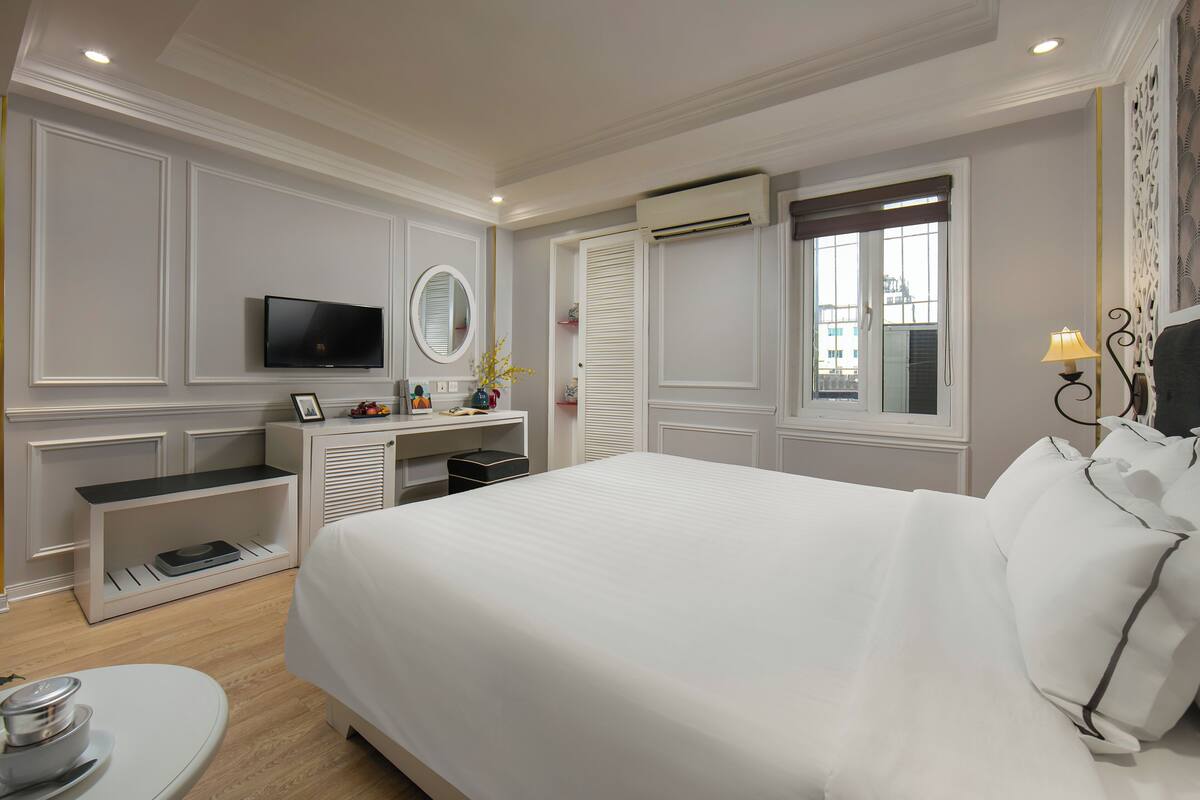 A modern hotel room with a large bed, a TV, a vanity with a mirror, a desk, and a window with shutters.