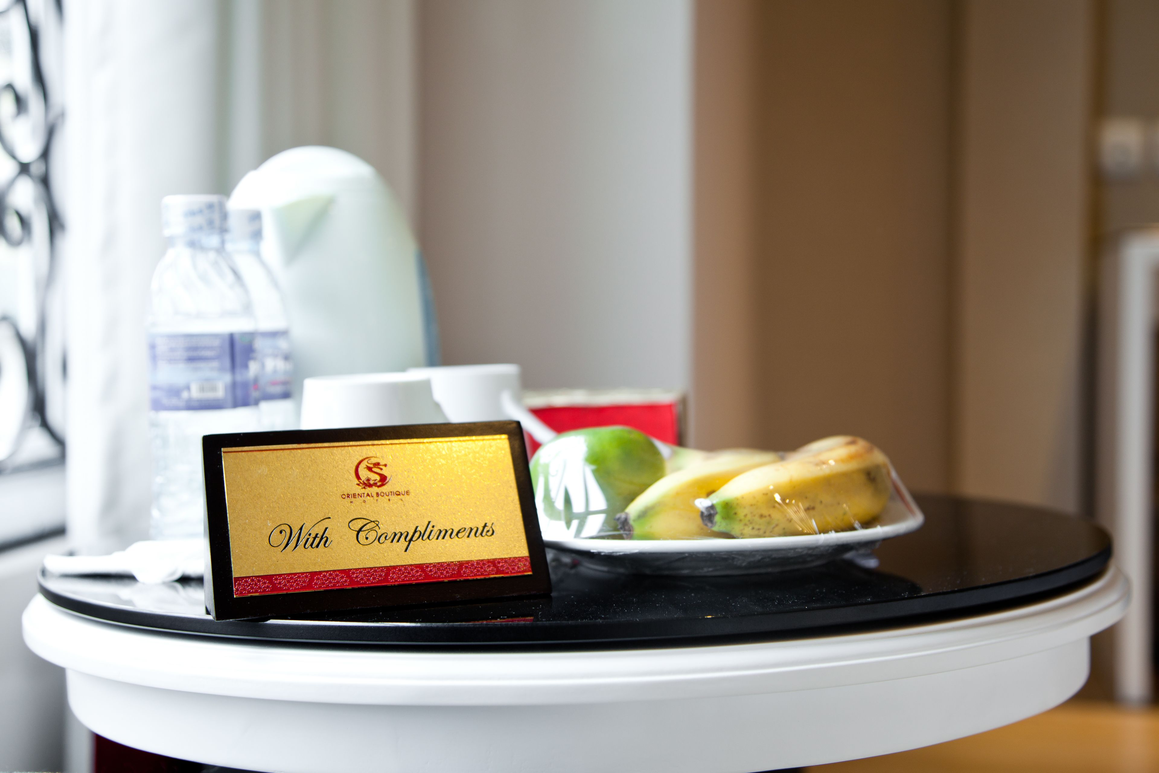Ein Tablett mit einem Schild, das 'Mit freundlichen Grüßen' sagt, einer Wasserflasche, einer weißen Tasse und einem Teller mit Bananen und Äpfeln.
