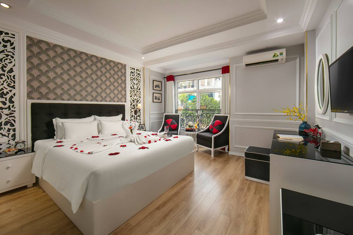 A modern hotel room with a large bed, two armchairs, a small table with a vase, and a TV.